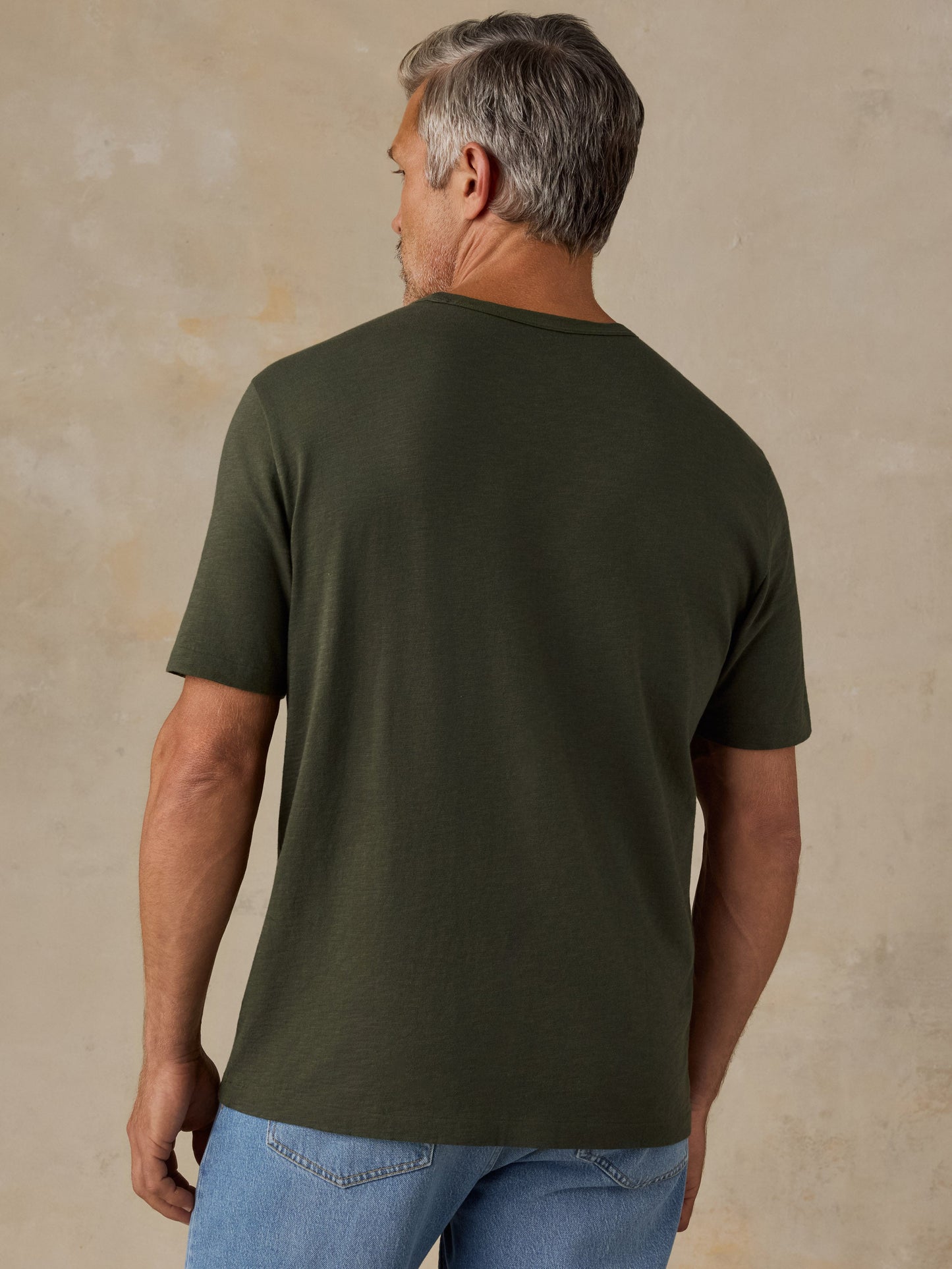 Male model wearing dark olive green Faherty Sunwashed Pocket Tee, back view with jeans