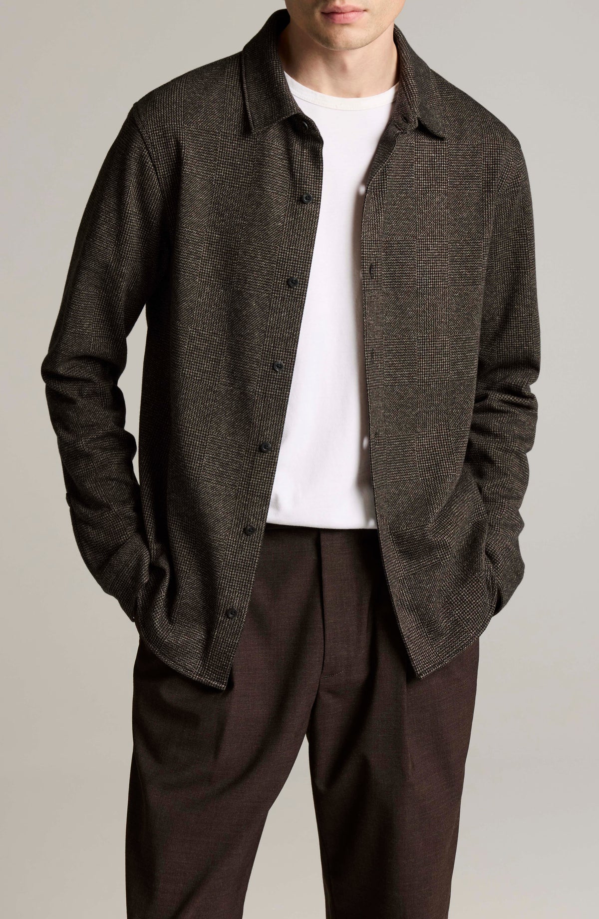 Male model wearing open dark brown glen plaid long sleeve button-down shirt over white tee with brown trousers