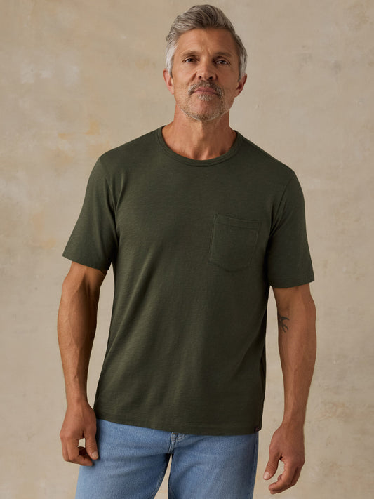 Man wearing Faherty Sunwashed Pocket Tee in dark olive green with chest pocket and blue jeans
