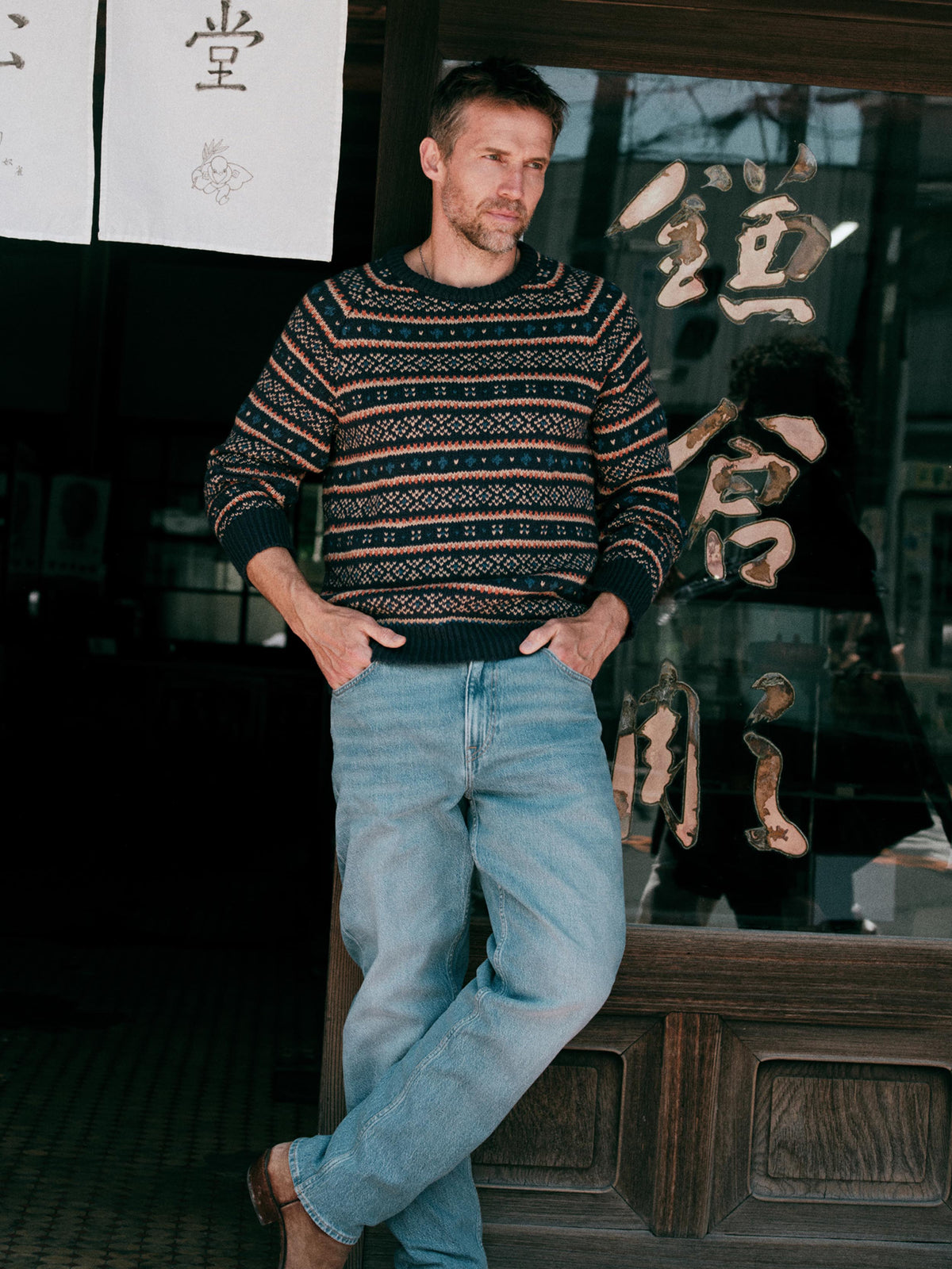 Male model wearing Faherty Nordic Sweater Crew in Northern Nights Fairisle with light wash jeans outside Japanese storefront