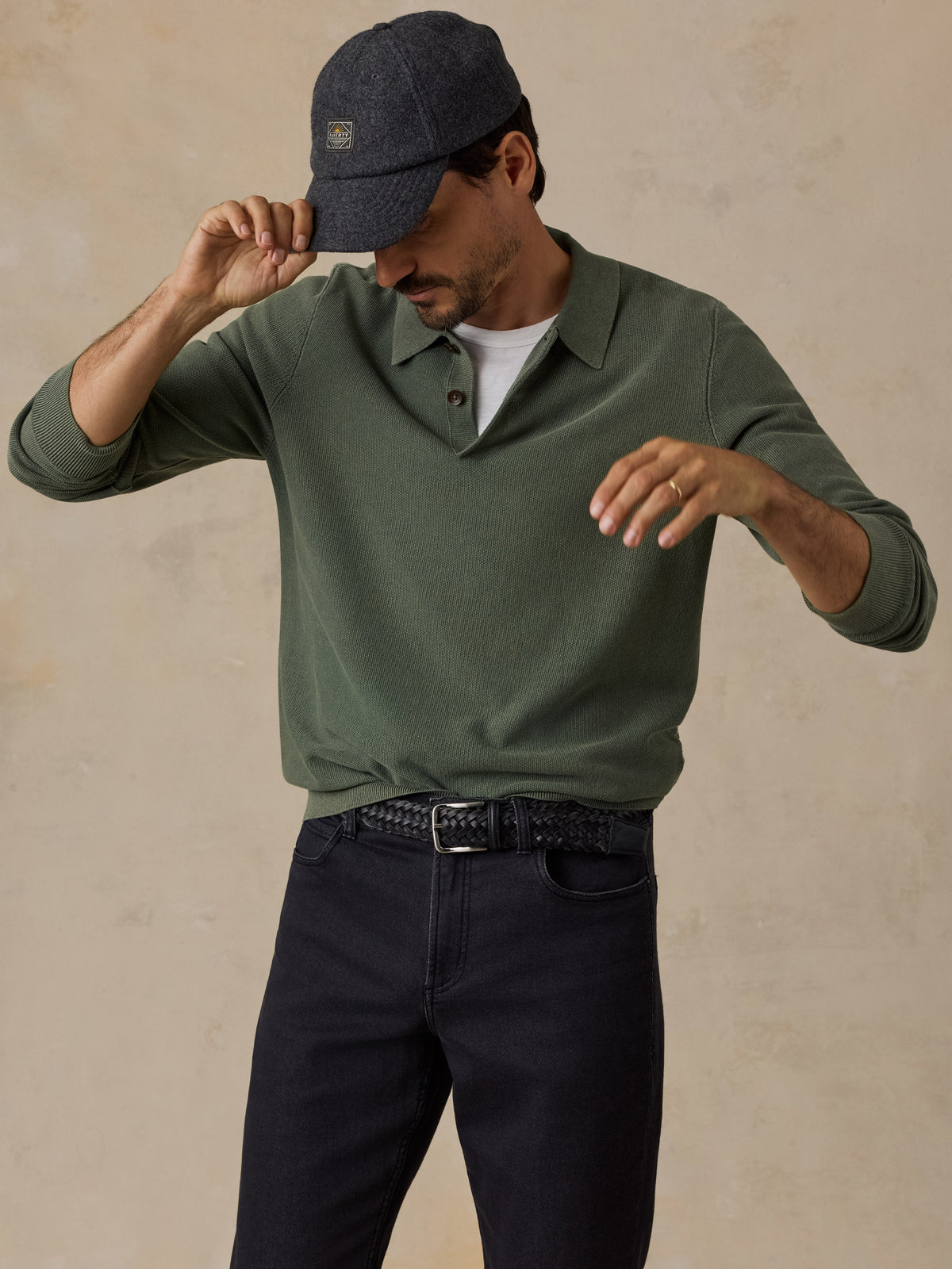 Male model wearing sage green long-sleeve sweater polo with dark jeans and grey cap