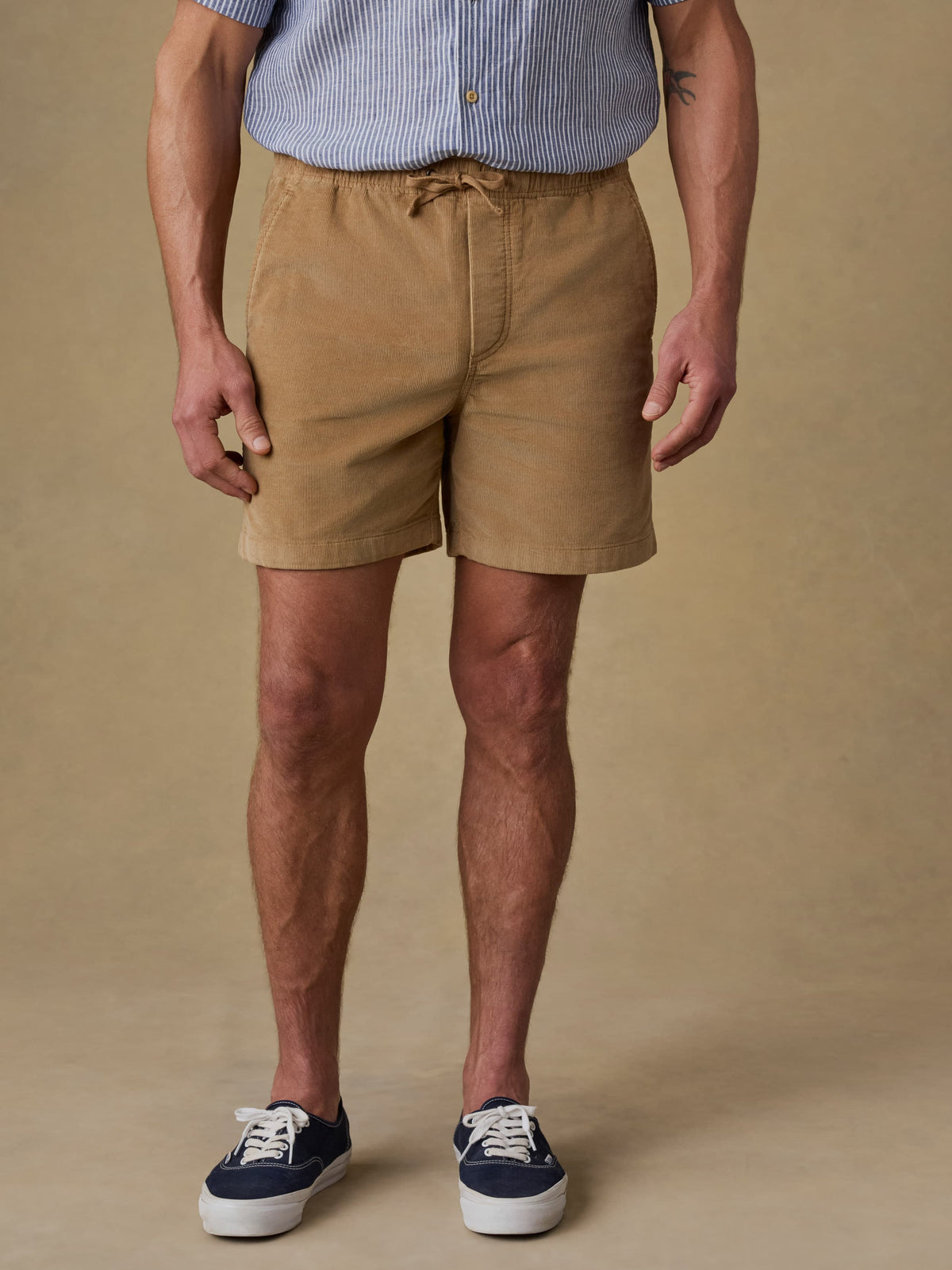 Man wearing tan corduroy shorts with striped shirt and sneakers against beige background
