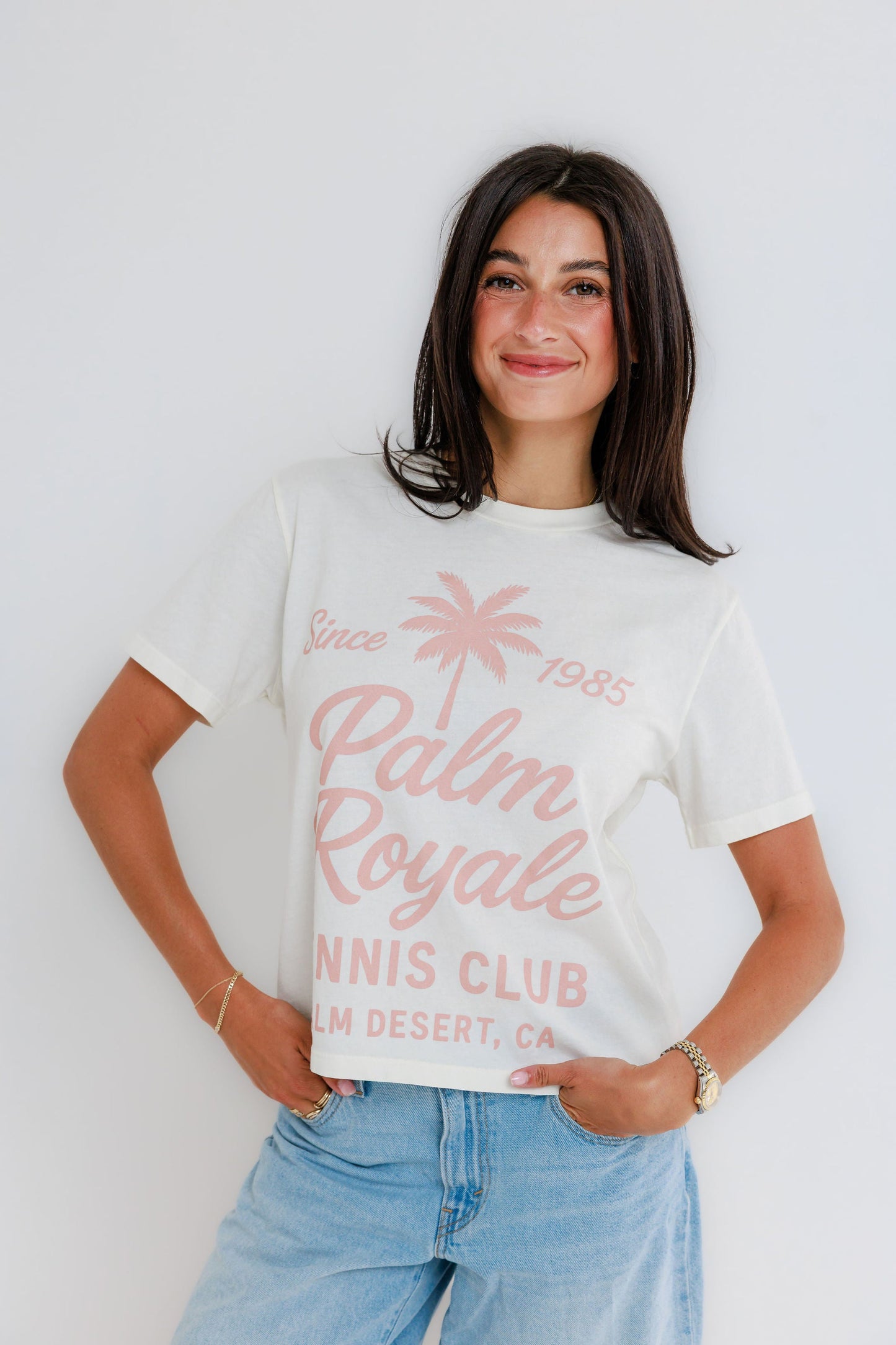 Woman smiling wearing cream Palm Royale Tennis Club graphic tee with pink print and jeans