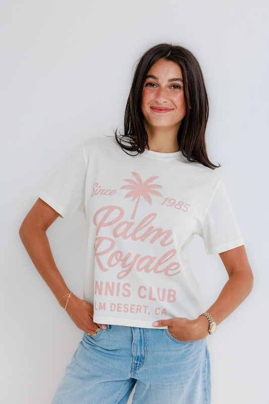 Woman smiling wearing cream Palm Royale Tennis Club graphic tee with pink print and jeans