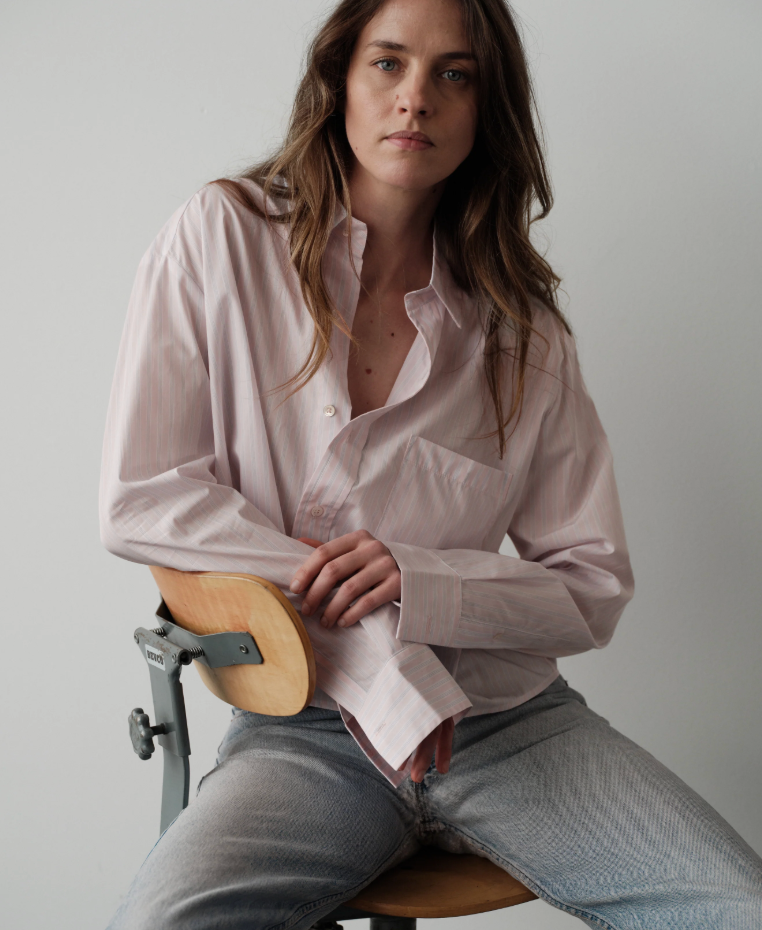 Woman wearing Cissa oversized button-up shirt in soft striped pattern, sitting casually on wooden chair.
