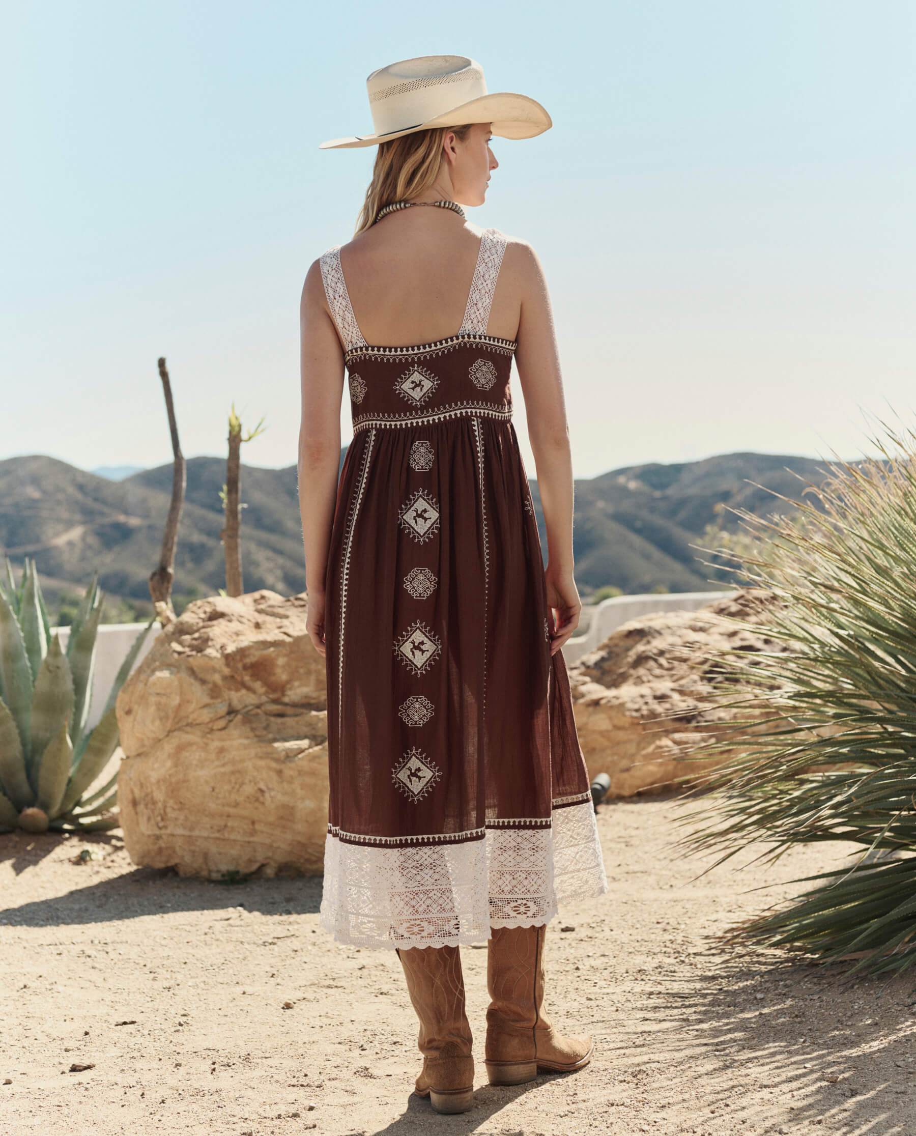 Model wearing chocolate brown embroidered midi dress with white lace trim, cowboy hat and boots in desert setting
