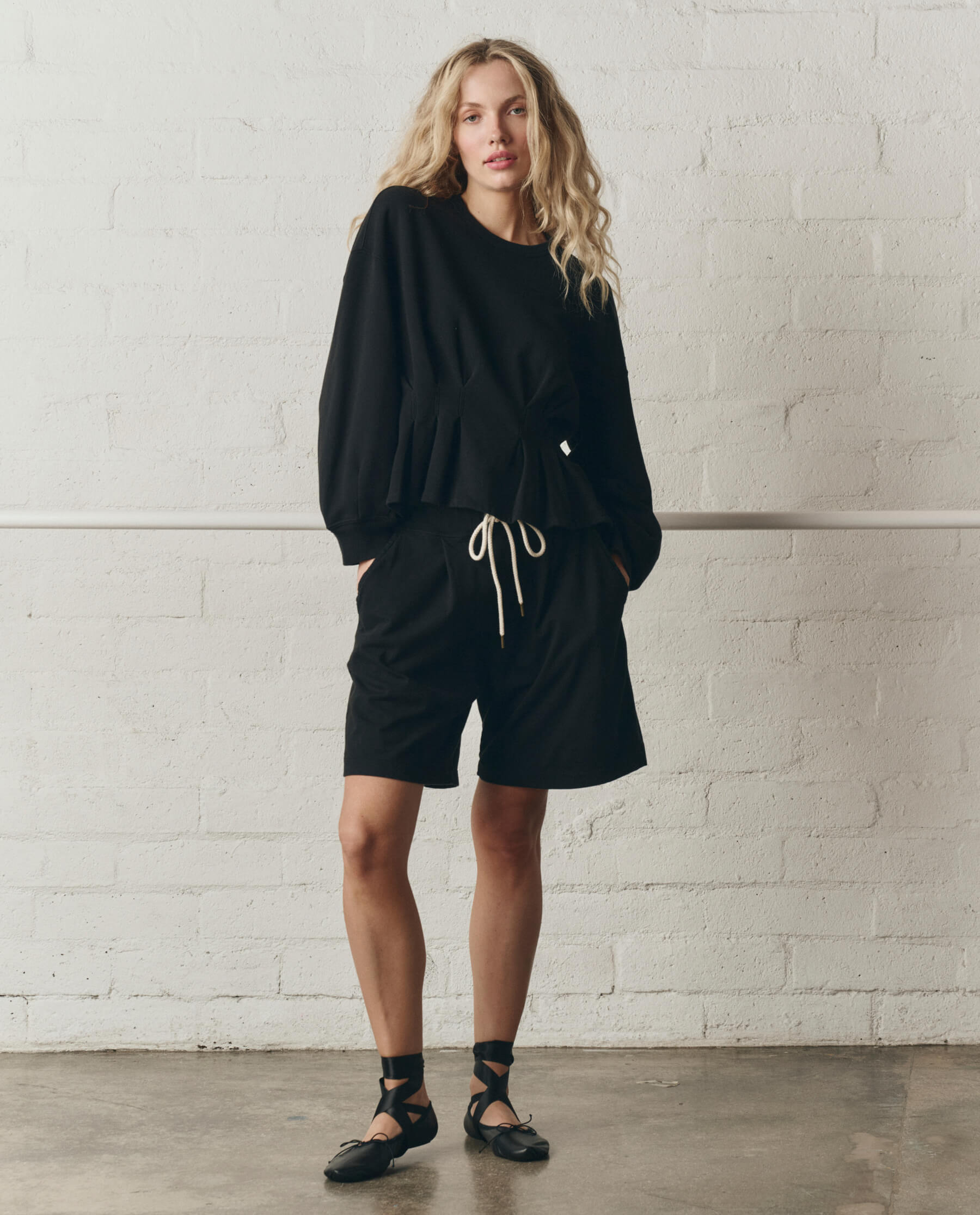 Woman wearing black corset sweatshirt and matching shorts against white brick wall