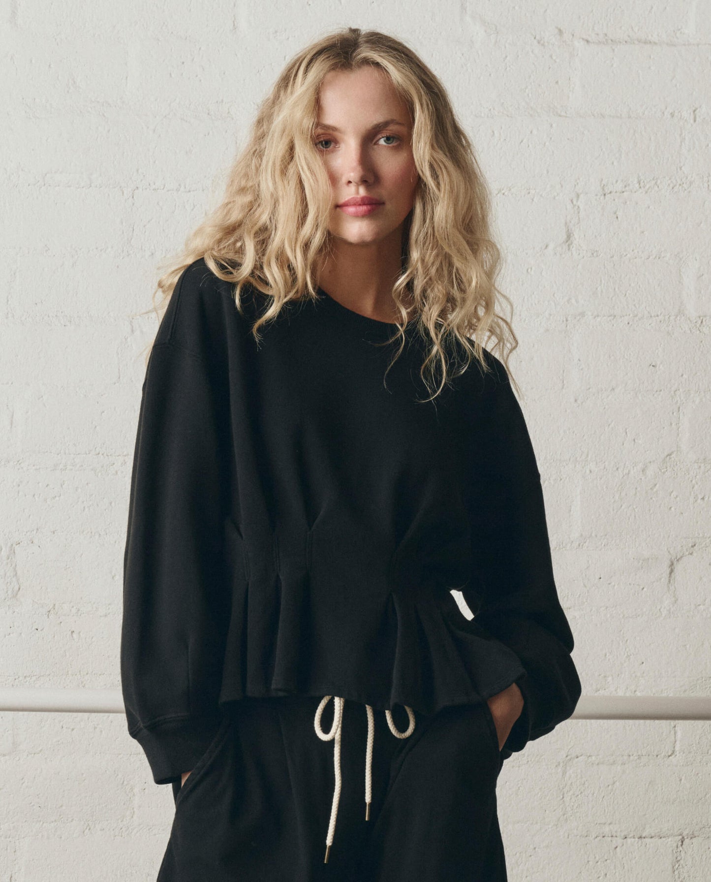 Woman wearing black slouchy corset sweatshirt with drawstring waist against white brick wall