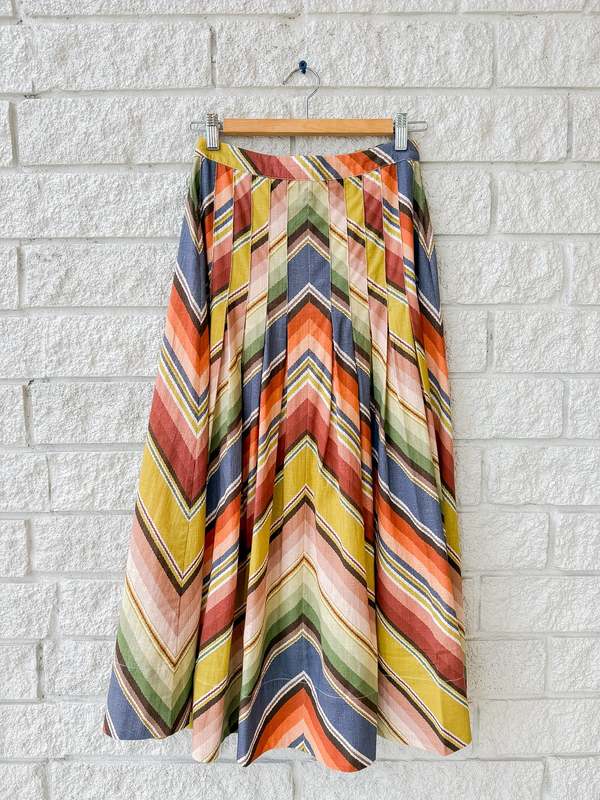 Multicolor chevron stripe midi skirt with pleats hanging on wooden hanger against white brick wall