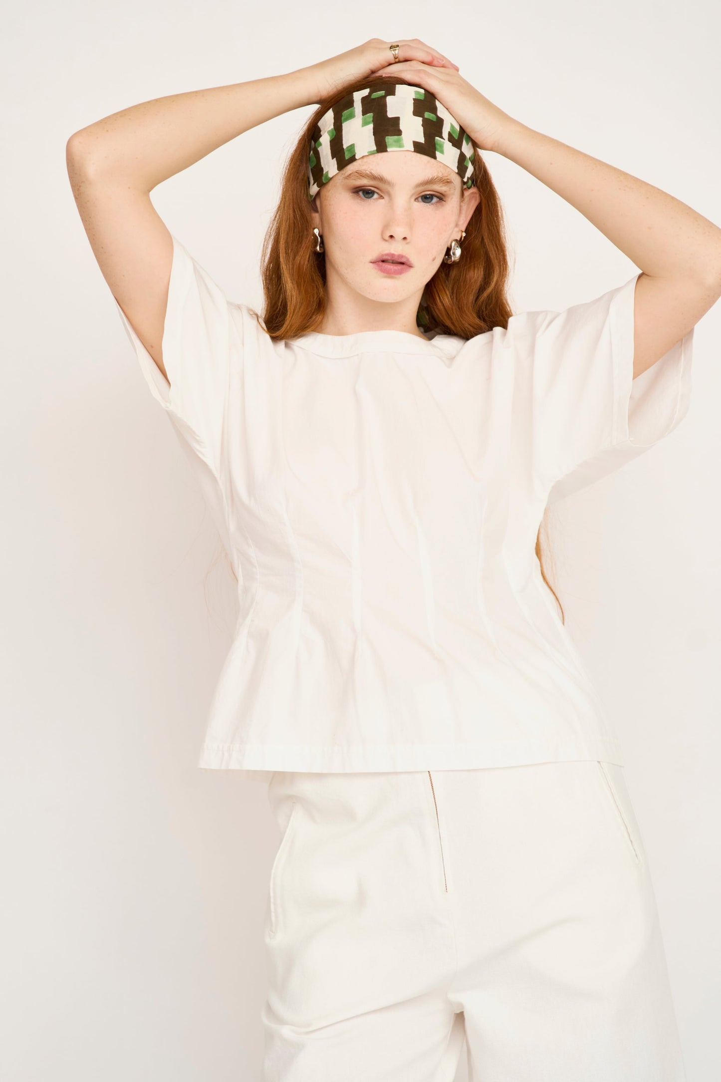 Woman wearing white oversized poplin top with headband, hands positioned above head