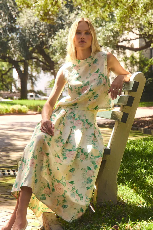 Woman wearing Hunter Bell Alexis floral midi skirt with matching sleeveless top, sitting on wooden bench in sunlit park se...