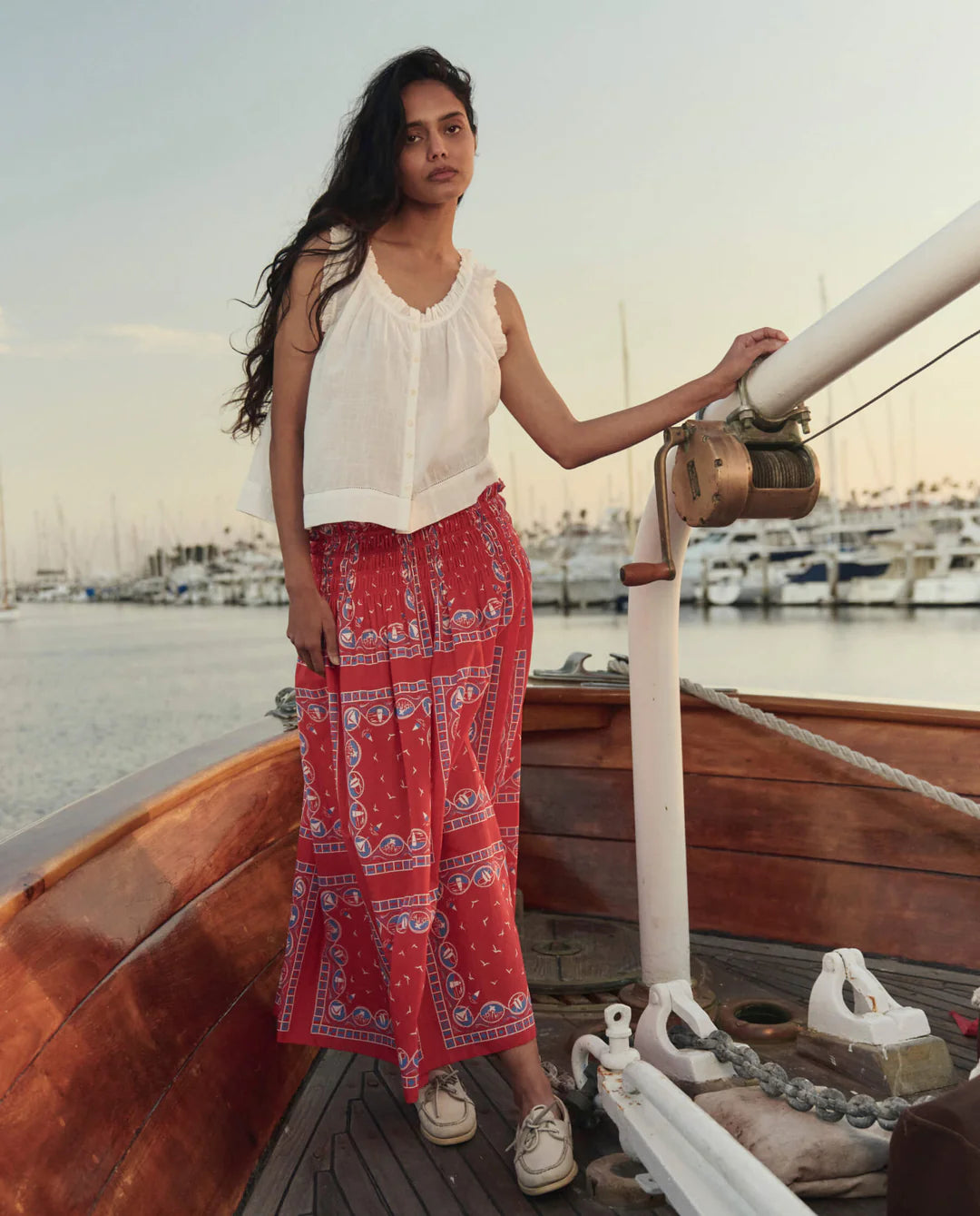 Woman wearing The Great red bandana print midi skirt with white sleeveless top on wooden boat deck.
