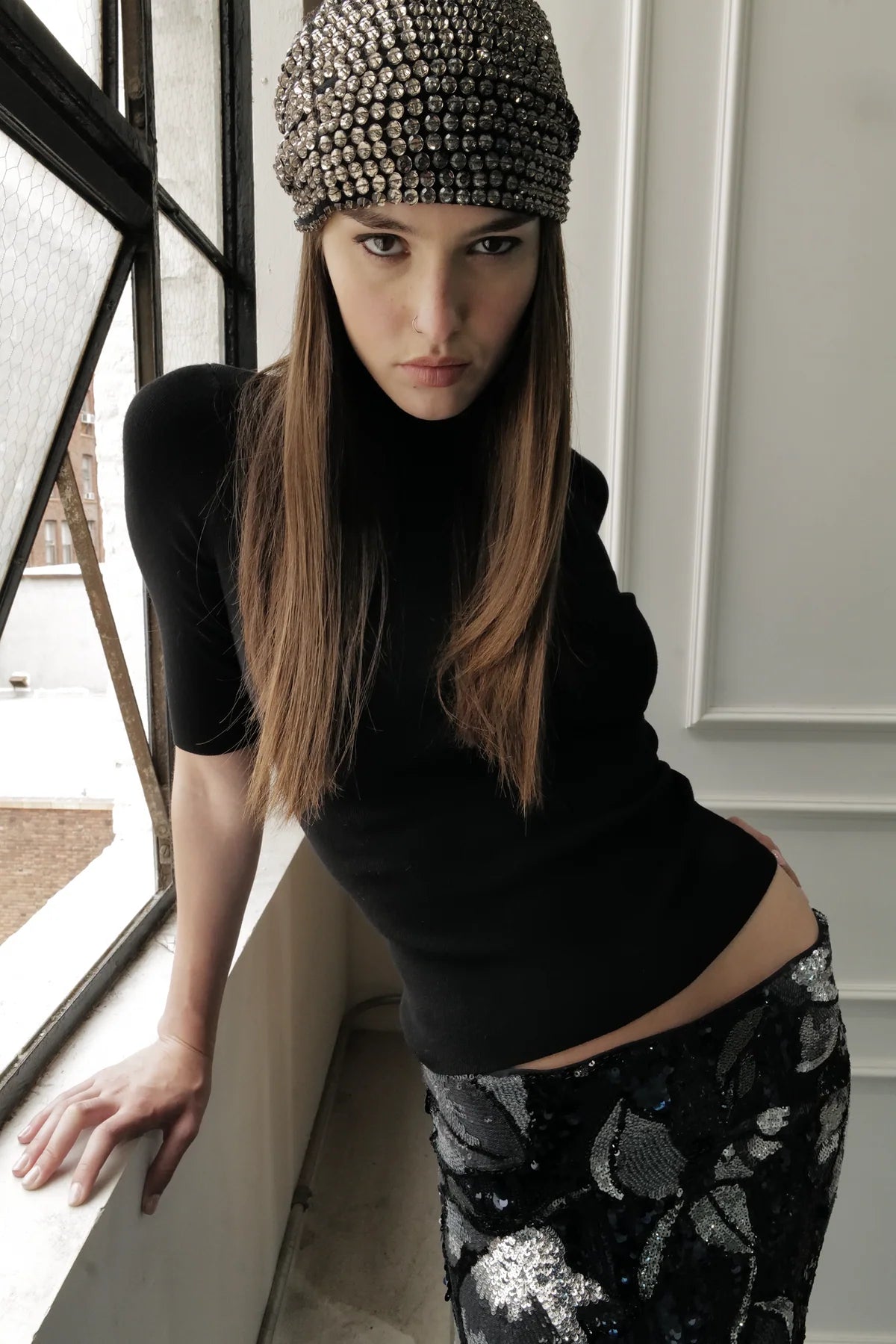 Woman wearing Le Superbe black knit top with studded beanie and sequined skirt in modern interior setting.
