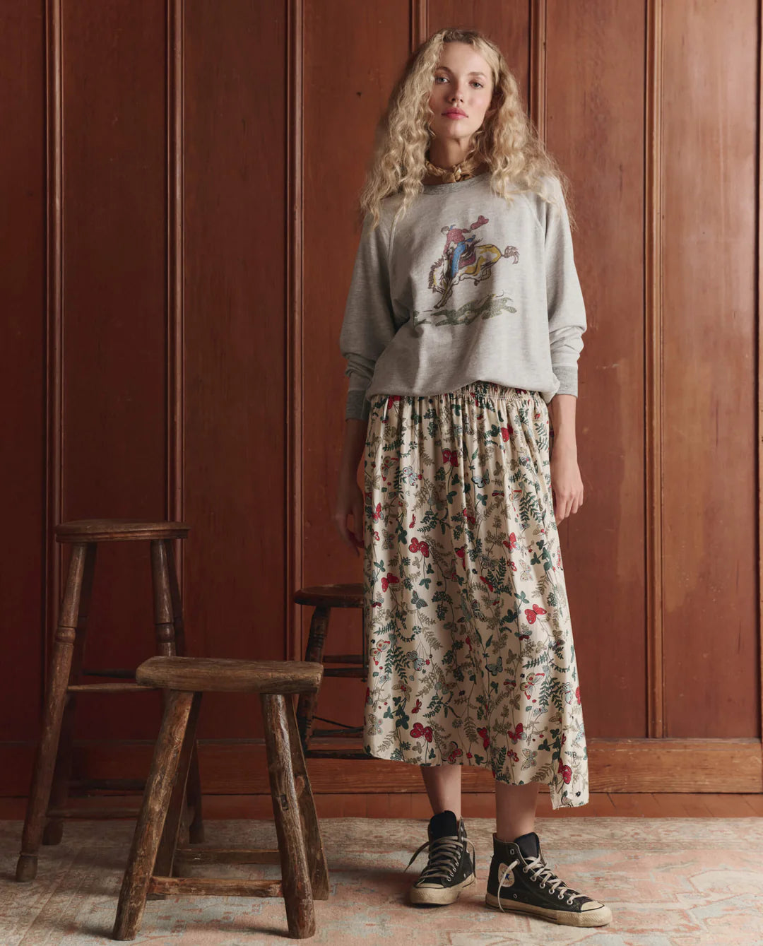 Woman wearing The GREAT floral midi skirt with elastic waist and black sneakers in rustic interior setting.