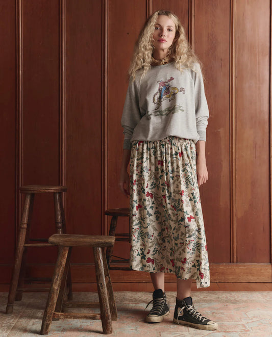 Woman wearing The GREAT floral midi skirt with elastic waist and black sneakers in rustic interior setting.