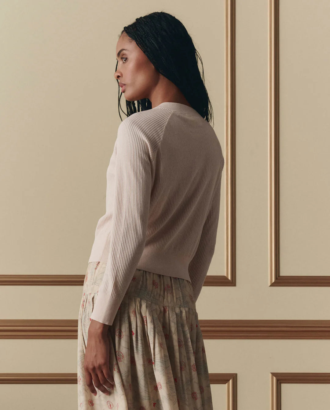 The Great ribbed cardigan sweater worn with patterned midi skirt, shown from back angle in elegant interior setting.