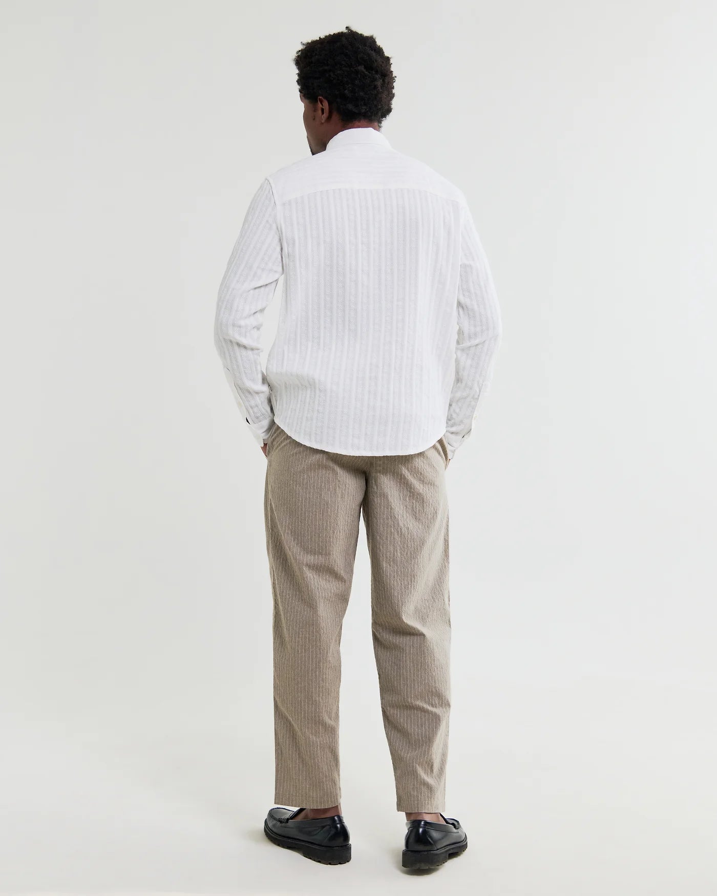 Back view of man wearing Wax London white striped long-sleeve shirt with beige trousers and black loafers.