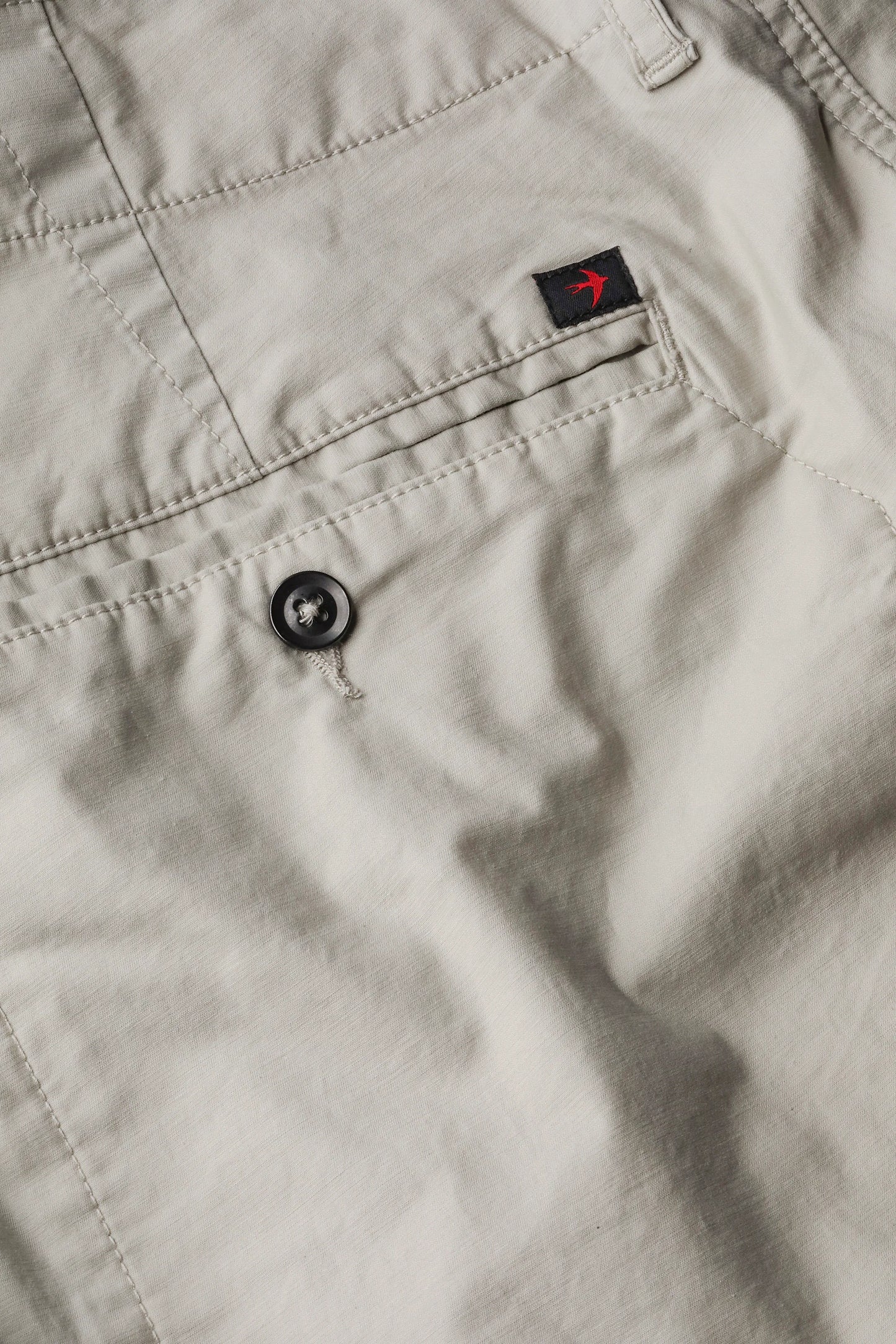 Relwen Flywt Flex chino pants detail showing front pocket, black button, and small branded label with red logo.