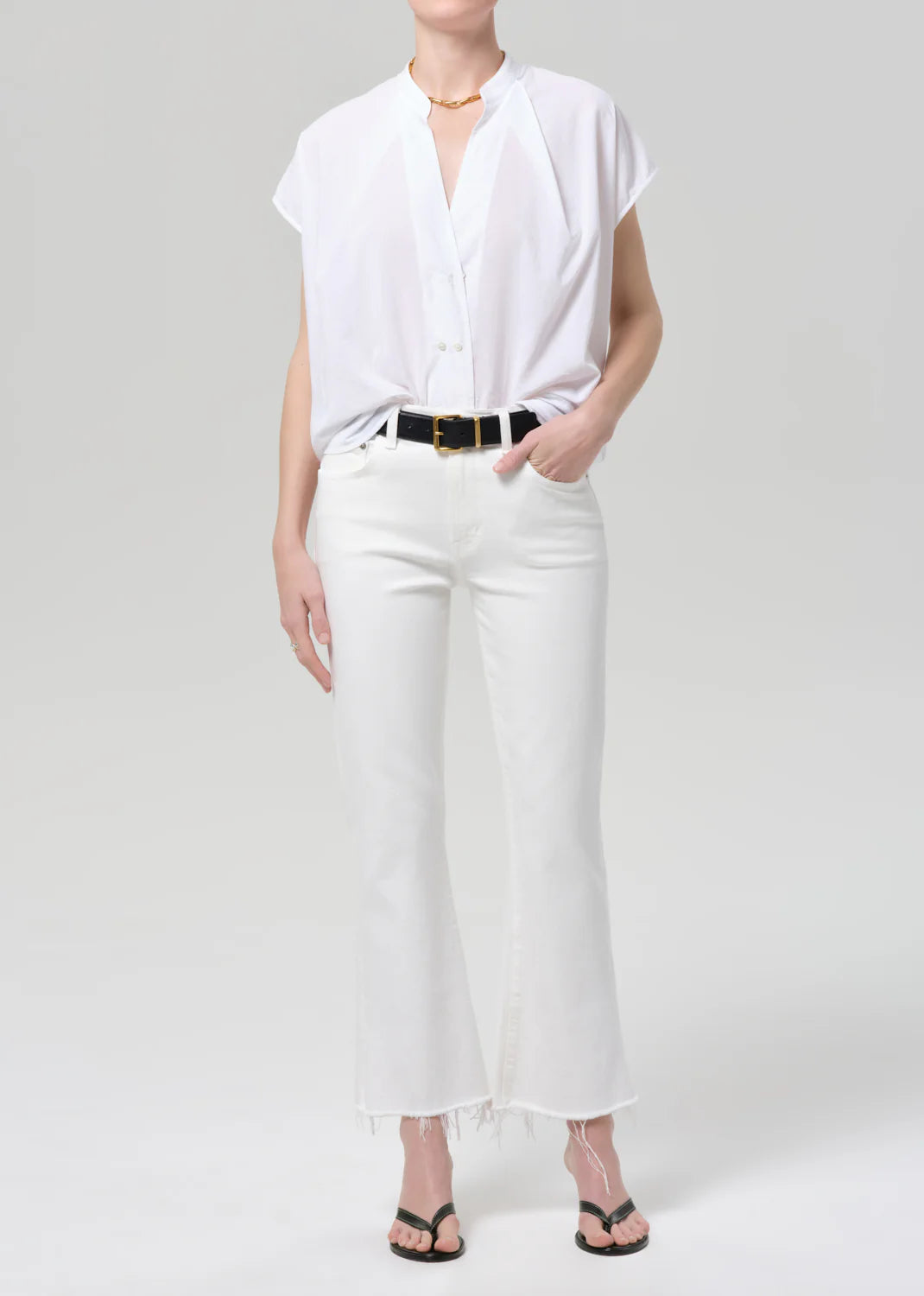 Woman wearing Citizens of Humanity white kick flare jeans with raw hem, white button-up shirt, and black sandals.