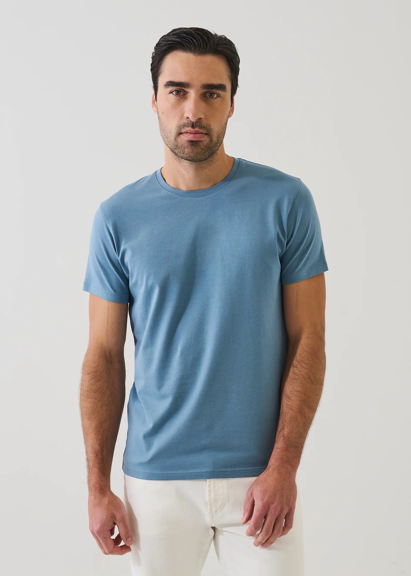 Man wearing Patrick Assaraf short sleeve crew neck t-shirt in haze blue with white pants against neutral background.