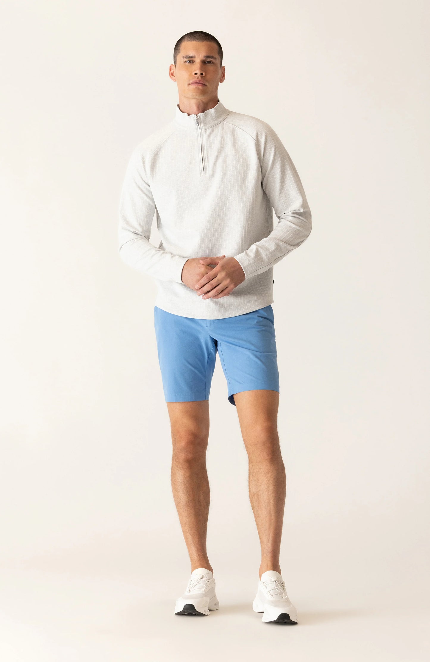 Man wearing white quarter-zip pullover and blue Deke Galaxy Bermuda shorts in Ultra 4X Stretch Twill, standing against neu...