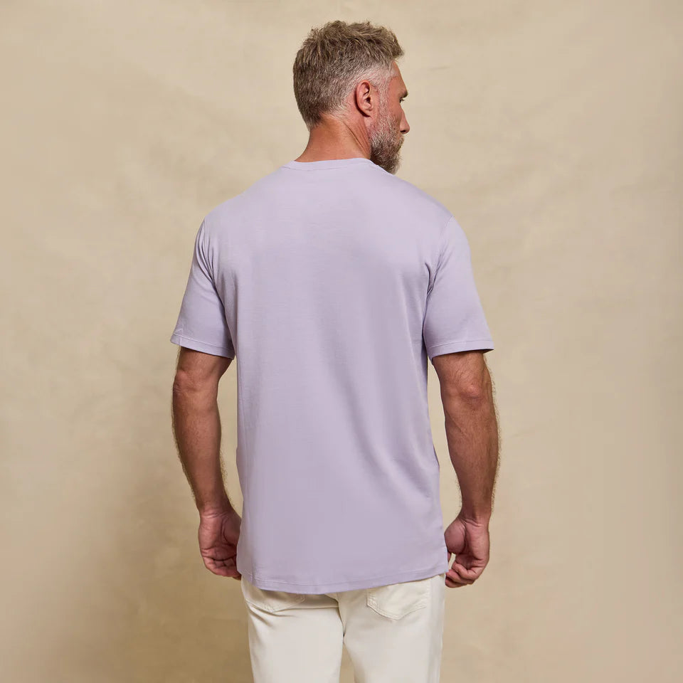 Back view of man wearing Rye 51 lavender short-sleeve crew neck t-shirt with cream pants against beige background.