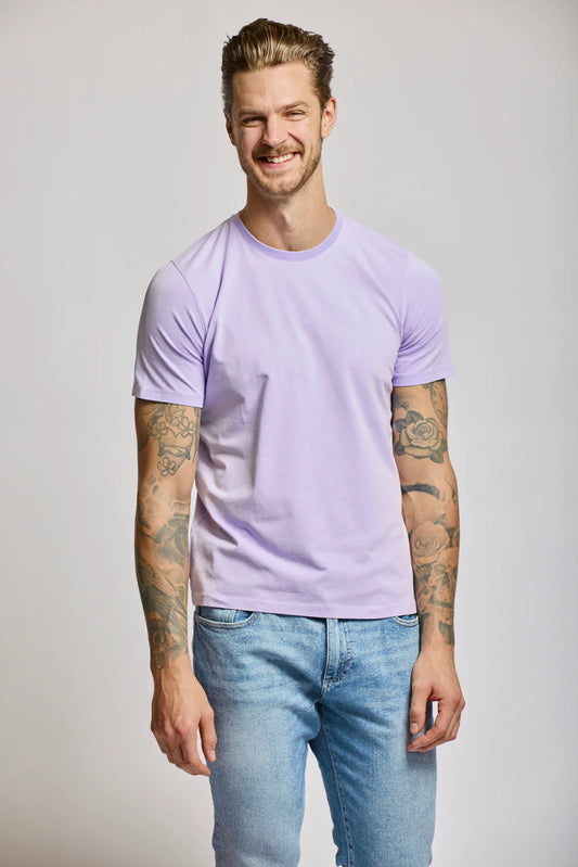 Man wearing Easy Mondays lilac crew neck t-shirt with blue jeans, showing tattooed arms and smiling.