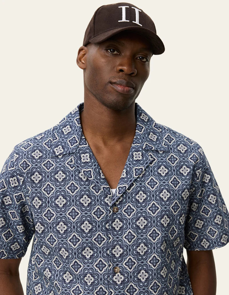 Man wearing Les Deux short-sleeve button-up shirt with blue geometric arabesque pattern and brown baseball cap.