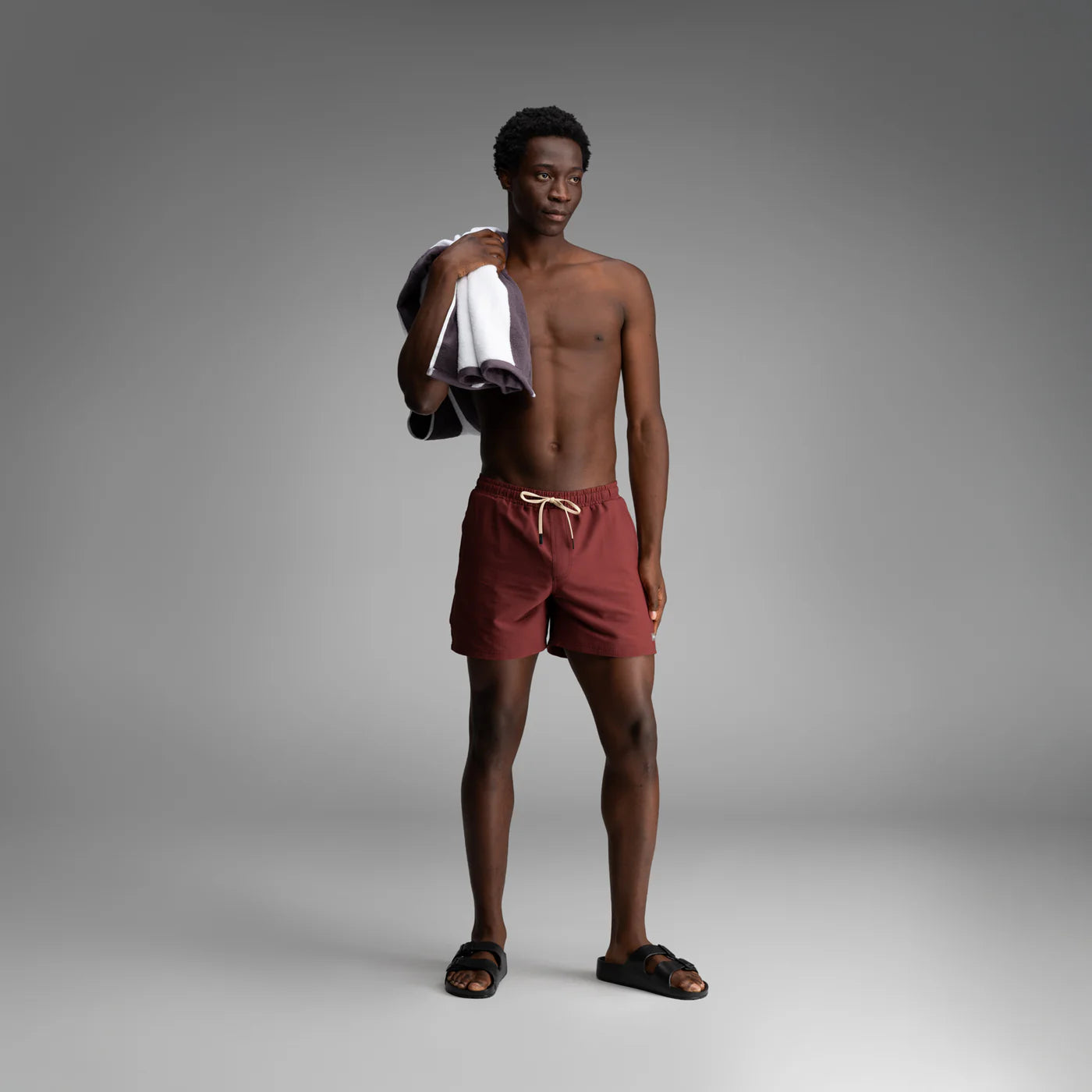 Man wearing BN3TH red swim trunks with drawstring waist, holding towel over shoulder in studio setting.