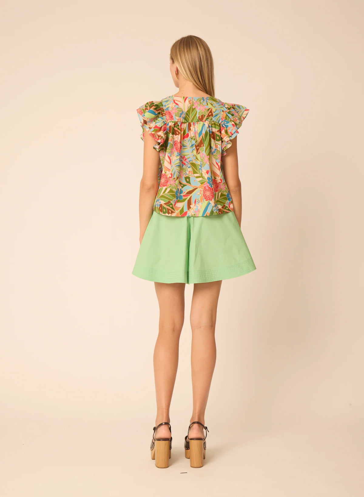 Back view of woman wearing Gilner Farrar Orla blouse with tropical floral print and ruffled sleeves, paired with green sho...