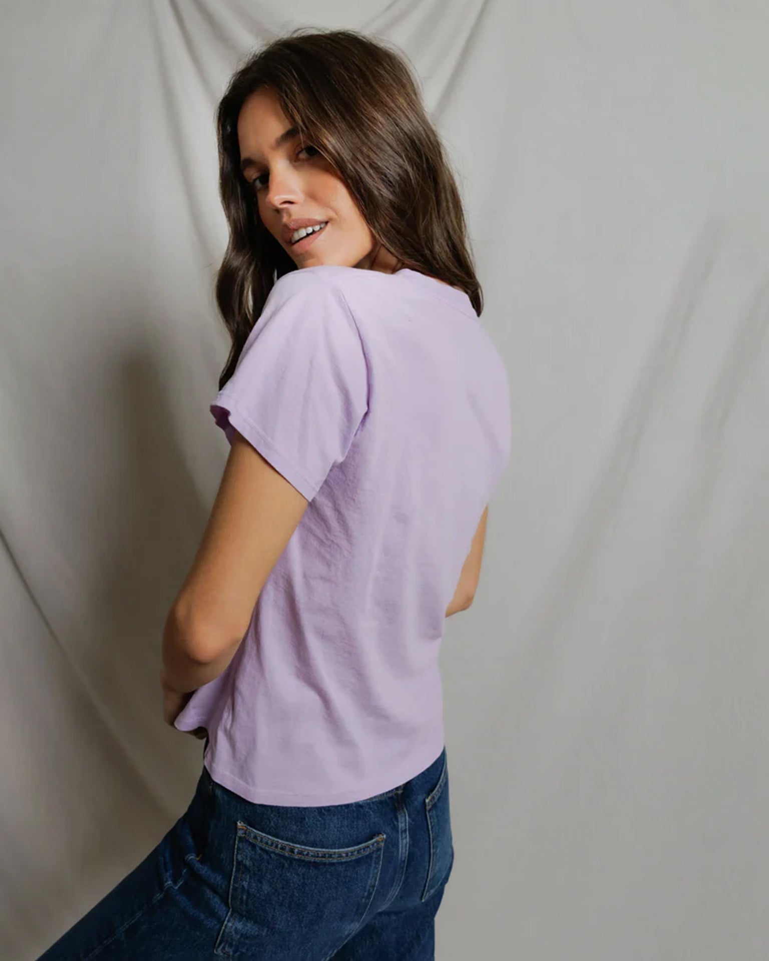 Woman wearing light purple v-neck short sleeve tee with jeans looking over shoulder