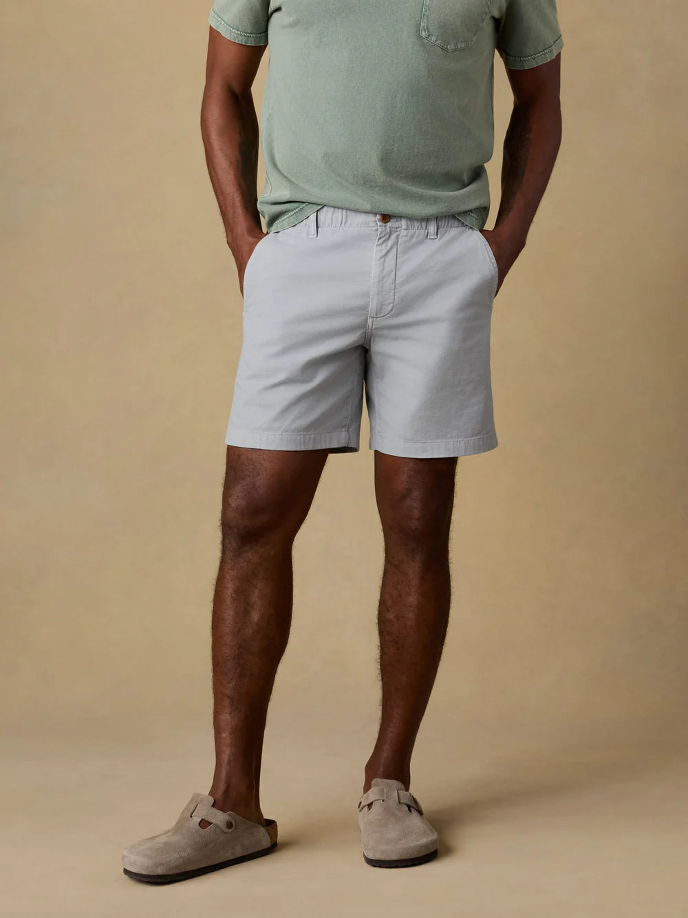 Man wearing Faherty Essential Chino Shorts in grey with green t-shirt and suede clogs on neutral background.