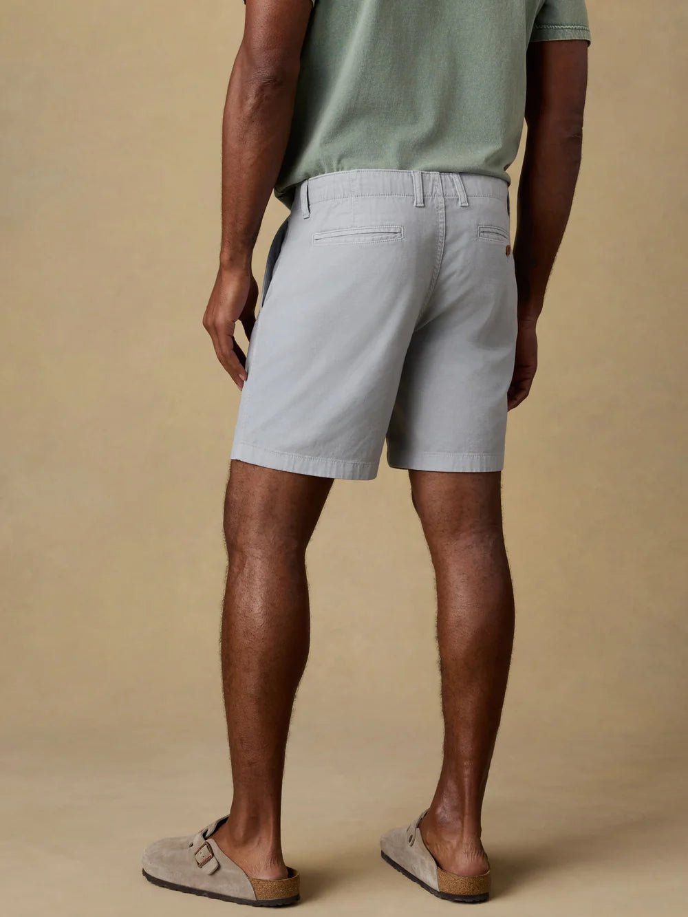 Man wearing Faherty Essential Chino Shorts in grey, shown from behind with casual sandals and green t-shirt.