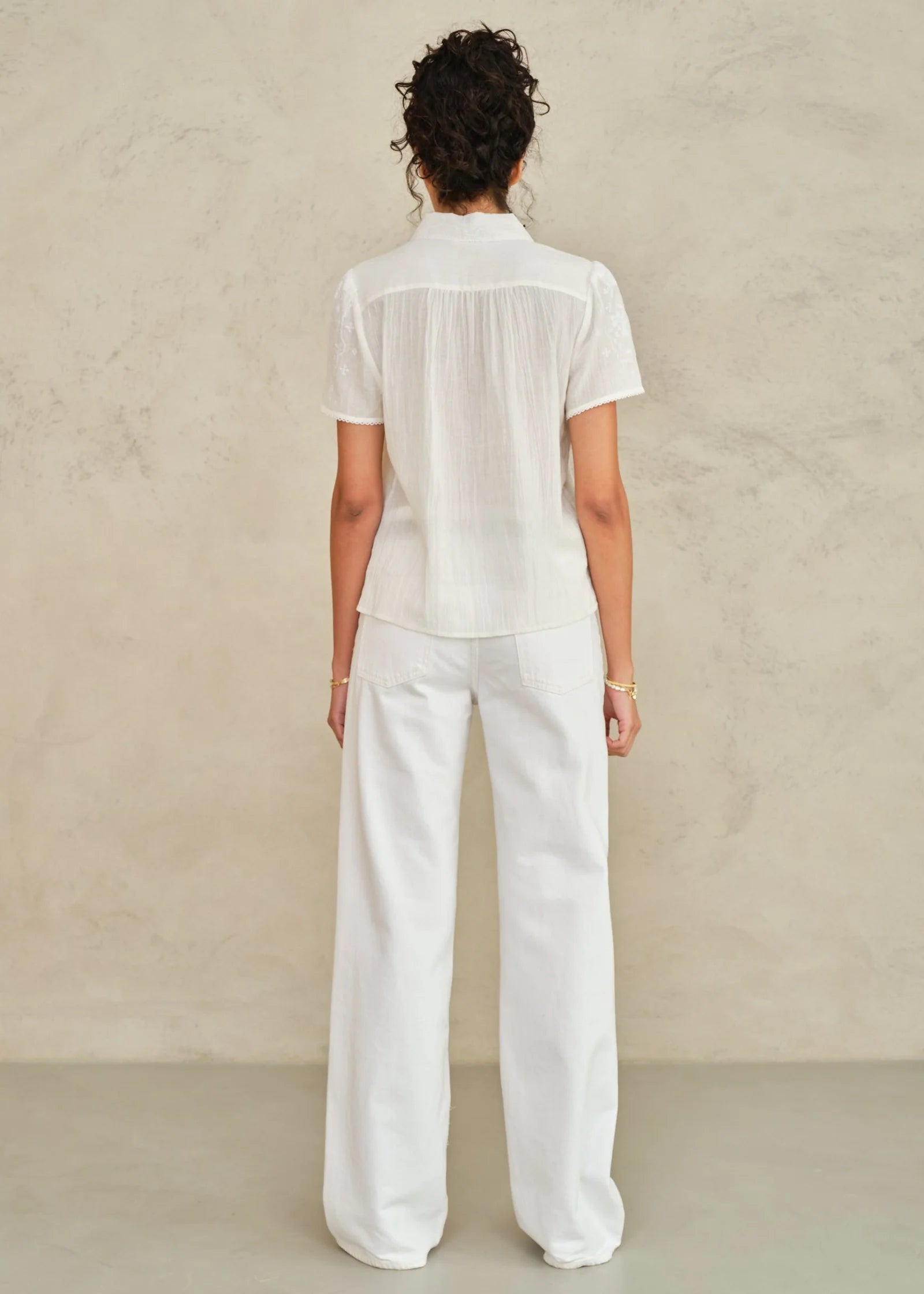 Back view of woman wearing white short-sleeve embroidered top and matching wide-leg pants from MABE Ruby collection.