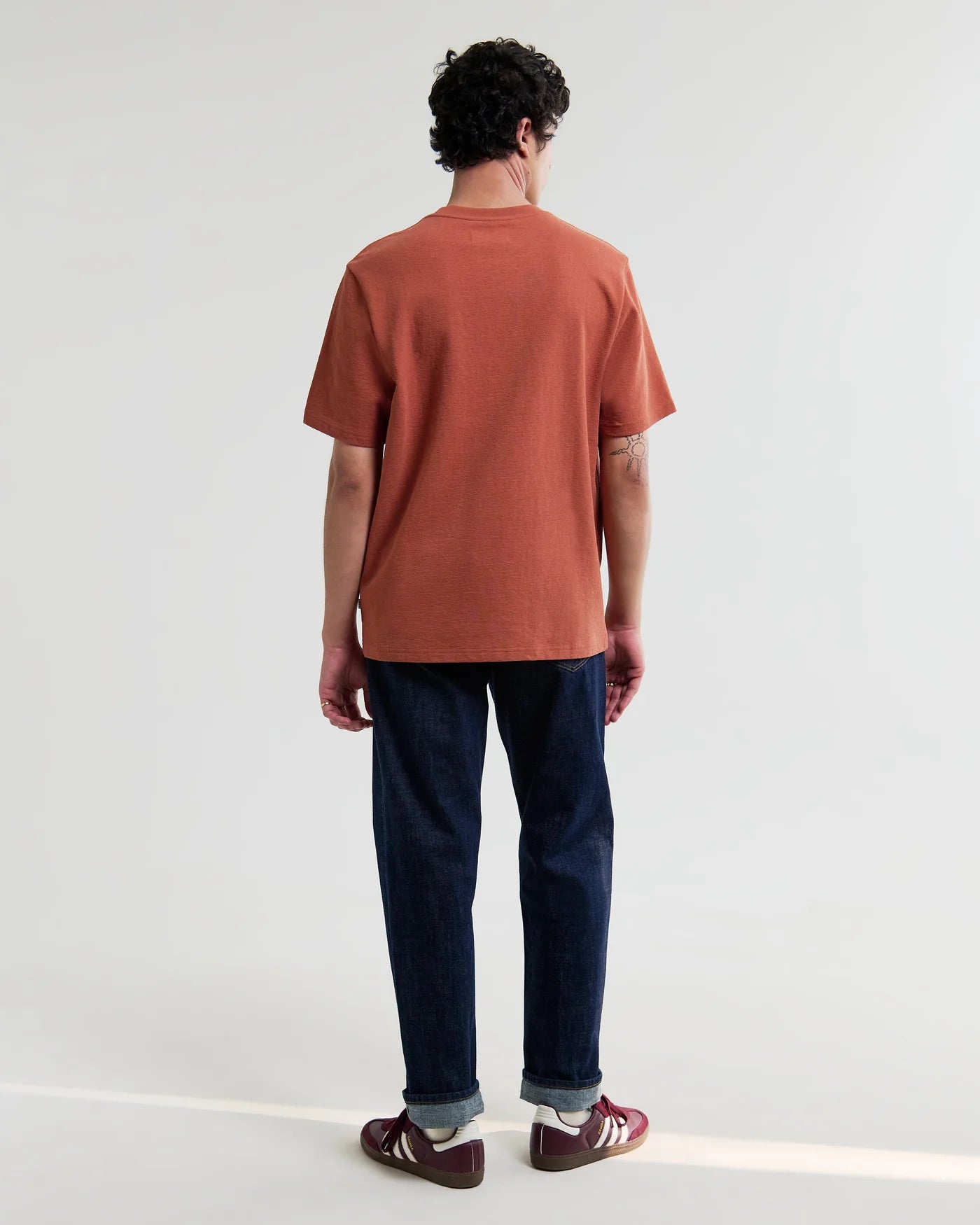 Back view of man wearing Wax London Dean short sleeve rust colored t-shirt with dark jeans and sneakers.
