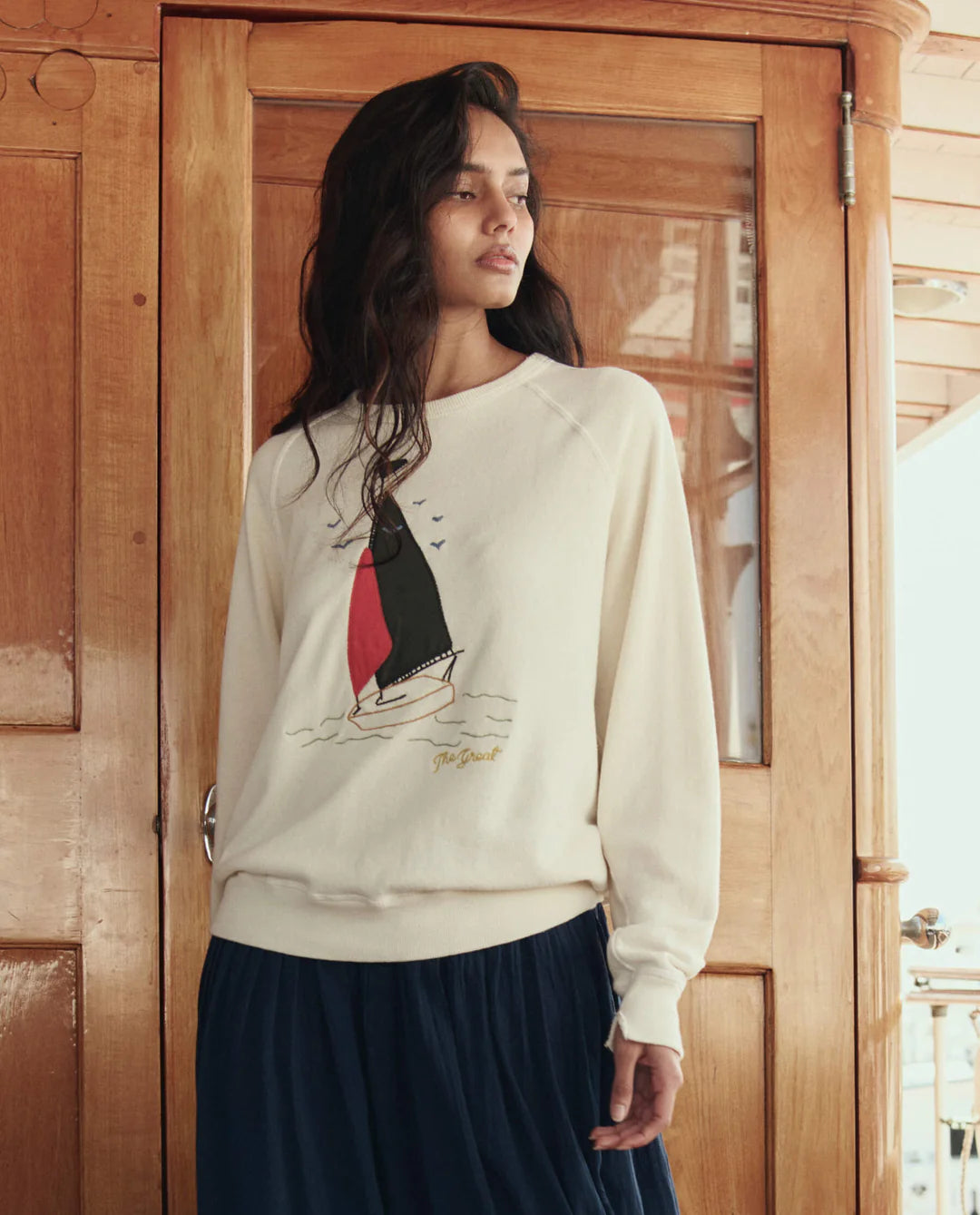 Woman wearing The GREAT College Sweatshirt with sailing boat applique design in cream, styled with navy skirt.