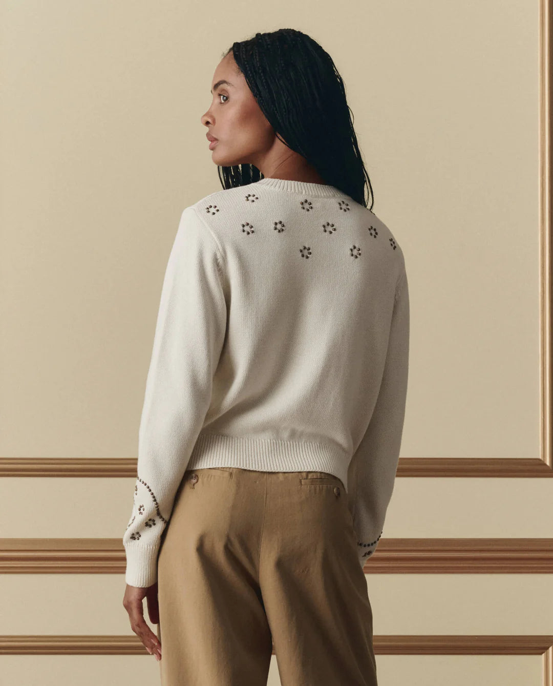 Woman wearing The Great studded cardigan in natural with decorative eyelet details on back and cuffs, paired with tan trou...