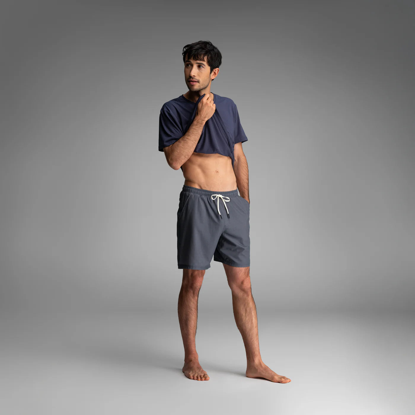 Man wearing BN3TH navy blue athletic shorts and matching t-shirt in casual pose against gray background.