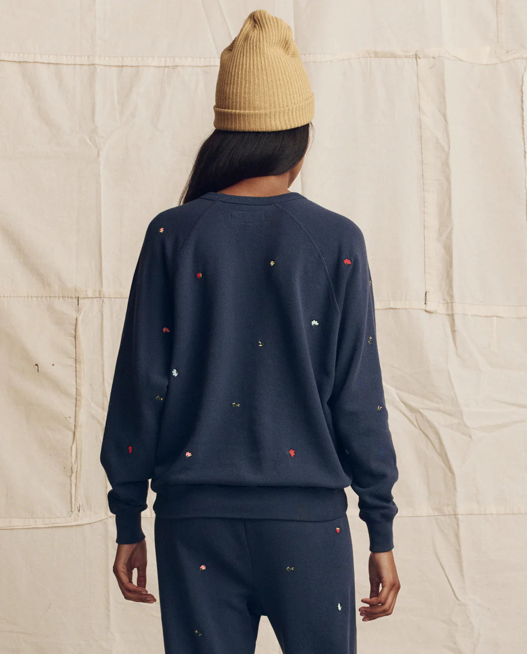 Woman wearing The Great navy sweatshirt with colorful floral embroidery and mustard knit beanie, viewed from behind.