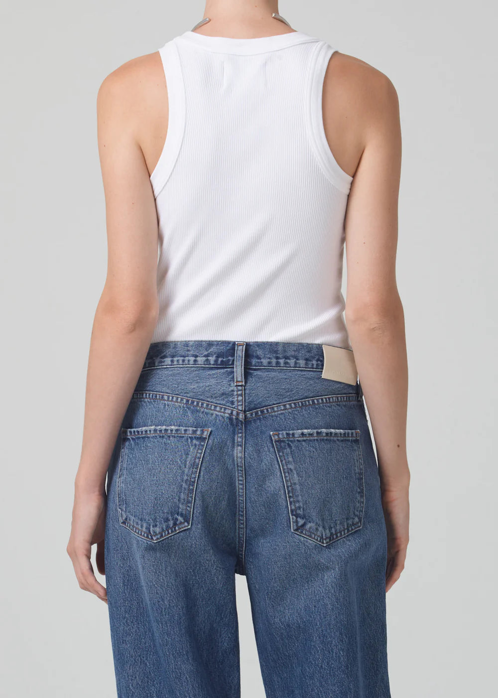 Back view of woman wearing Citizens of Humanity white ribbed tank top and blue jeans against gray background.